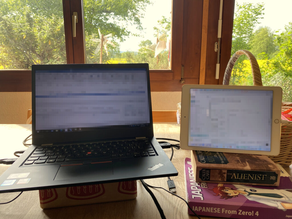Screenshot, der zeigt, wie ein iPad, auf Bücher gestützt, als erweiterter Bildschirm für ein Notebook verwendet wird. Der rechte Bildschirm (iPad) zeigt die erweiterte Anzeige des linken Notebooks. Das Setting ist improvisiert und entstand während eines Corona-Lockdowns.