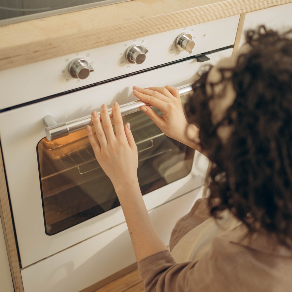 Frau kniet vor einem eingeschalteten Backofen in der Küche und hält ihre Hände davor, um die Wärme zu geniessen.