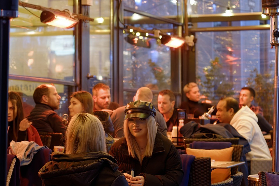 Gruppe von Menschen sitzt in einer Cafeteria, möglicherweise in einem Wintergarten. An der Decke sind Heizstrahler befestigt, die den Raum erwärmen.