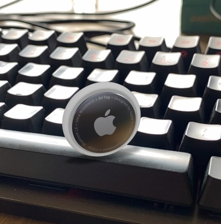 Ein Apple AirTag neben einer Computer-Tastatur, um den Grössenvergleich zu verdeutlichen. Der AirTag ist rund und silber, mit dem Apple-Logo, während die Tastatur ein modernes, schwarzes Design aufweist.